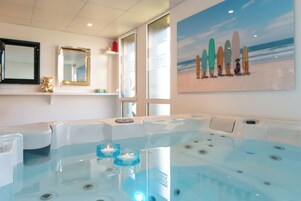 Uma área de spa com uma grande banheira de hidromassagem, um quadro com pranchas de surf na parede e um urso de pelúcia em uma estante.