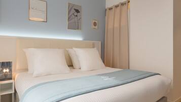 A neatly made bed with white linens and a light blue throw, a bedside table with a lamp, and two framed artworks on the wall.