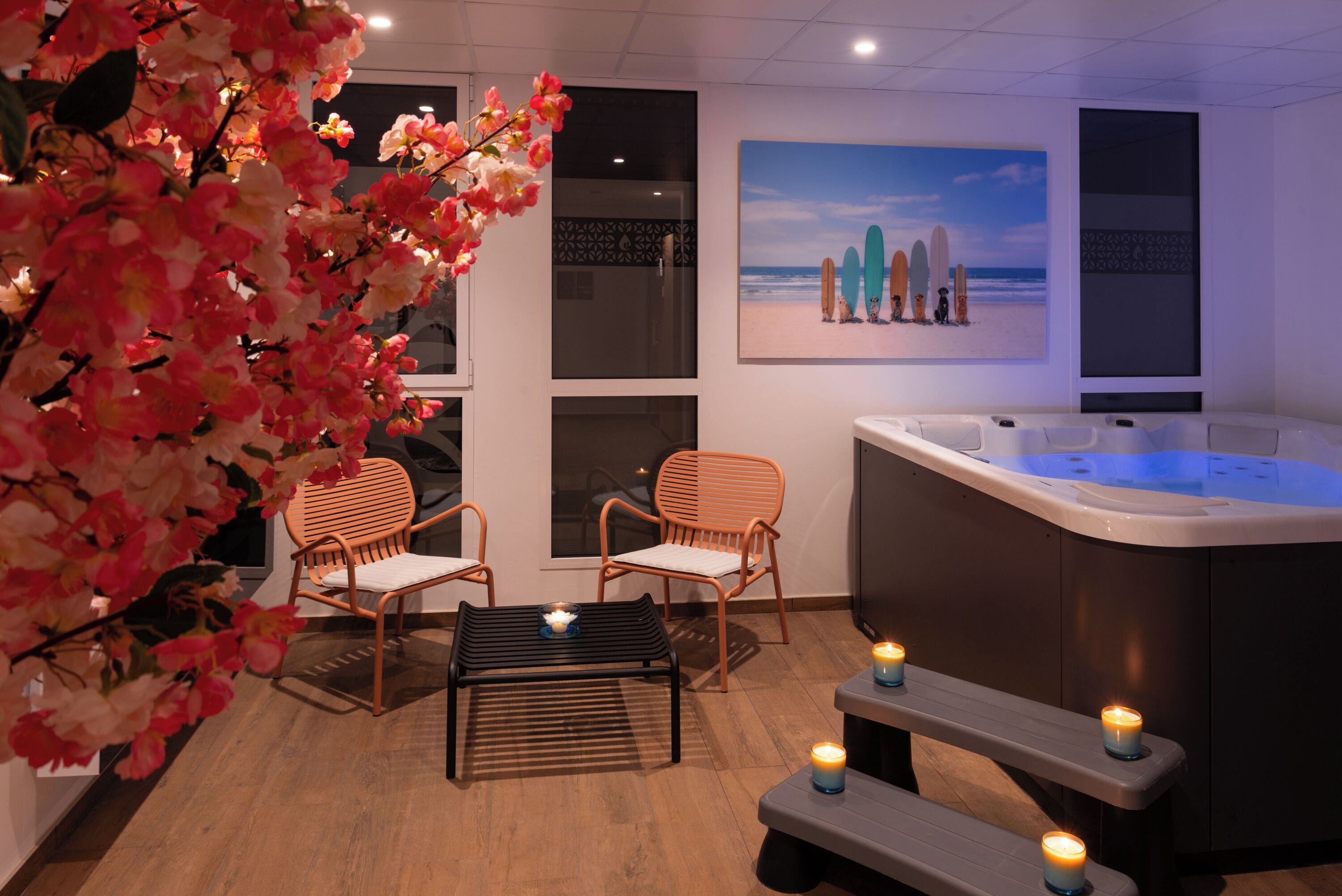 Ein Wellnessbereich mit Whirlpool, zwei Korbstühlen, einem Holztisch mit Kerzen und einem Wandbild im Strandhausstil.