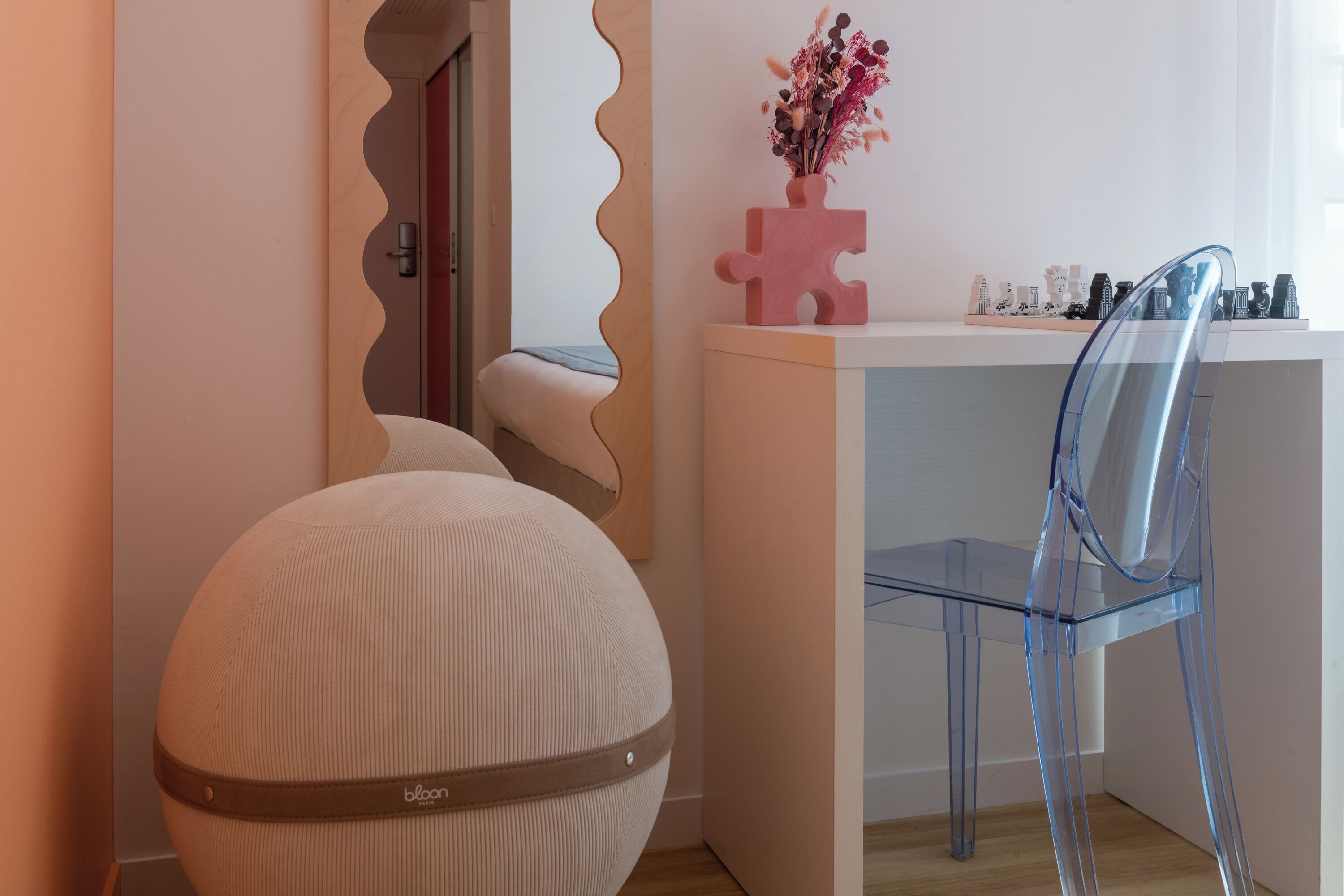 A room with a round ottoman, a clear chair, a white desk, and a mirror with a wavy frame.