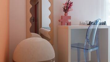 A room with a round ottoman, a clear chair, a white desk, and a mirror with a wavy frame.
