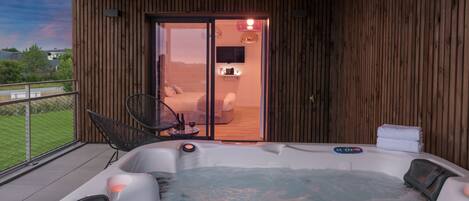 Un jacuzzi sur un balcon, avec un intérieur en bois, une chaise et une petite table sur laquelle sont posées des bouteilles.