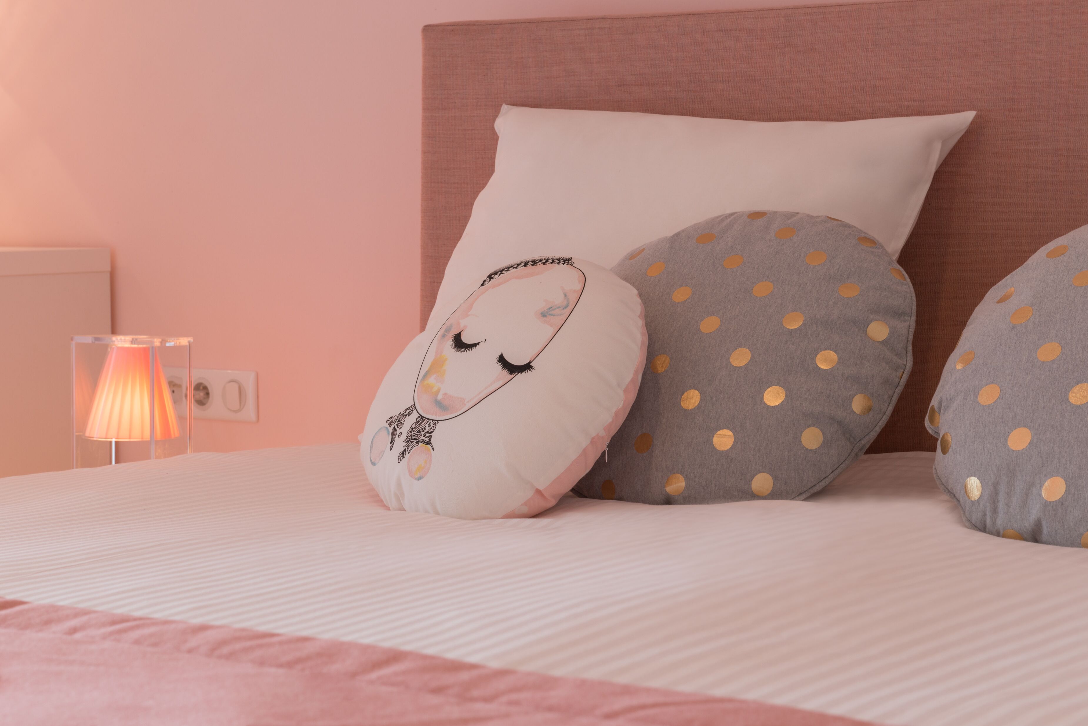 Un lit avec une couverture rose, un oreiller blanc orné d’un motif et deux oreillers gris à pois jaunes.