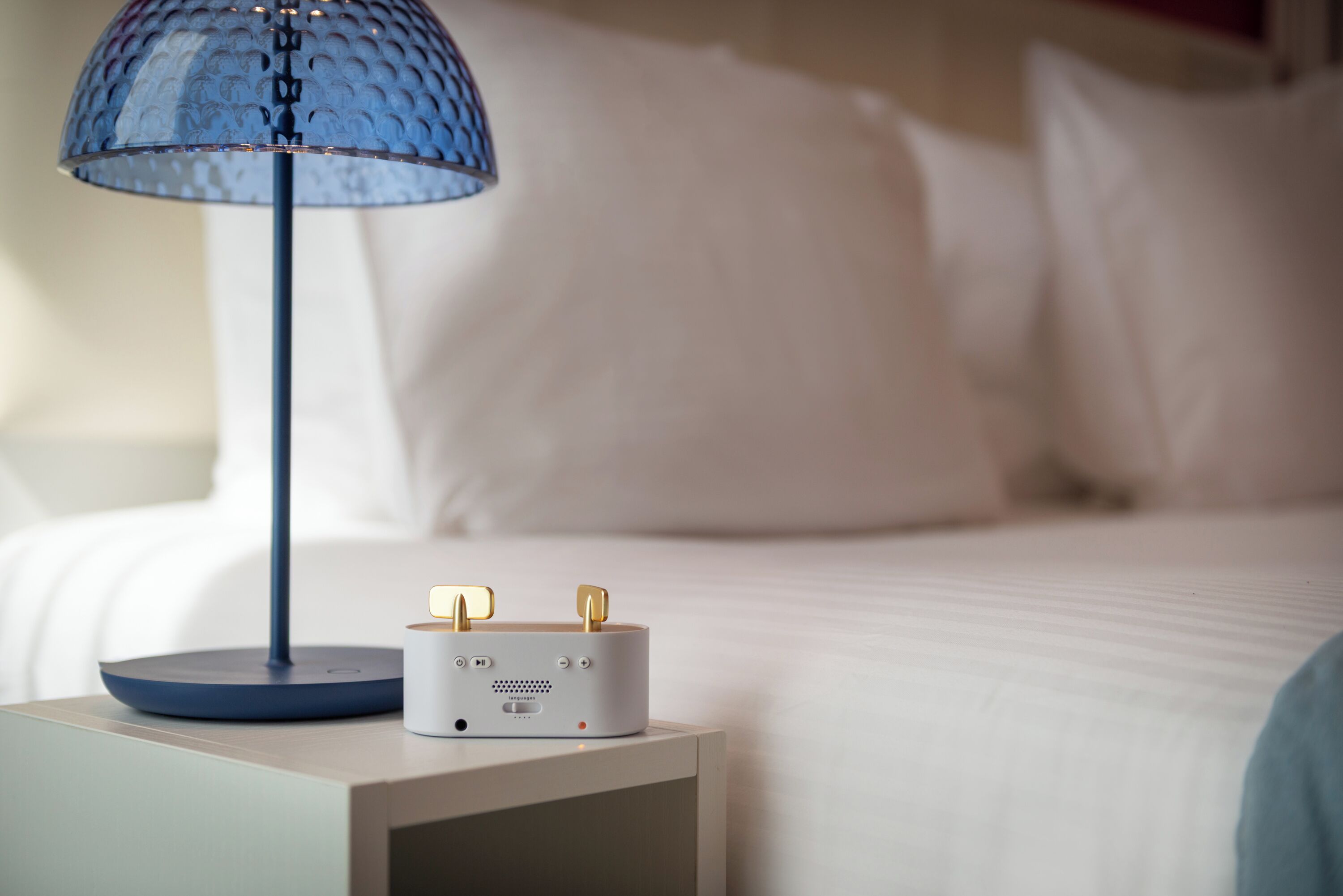 A bedside table with a lamp and a device, a bed with white bedding, and a wall in the background.