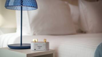 A bedside table with a lamp and a device, a bed with white bedding, and a wall in the background.