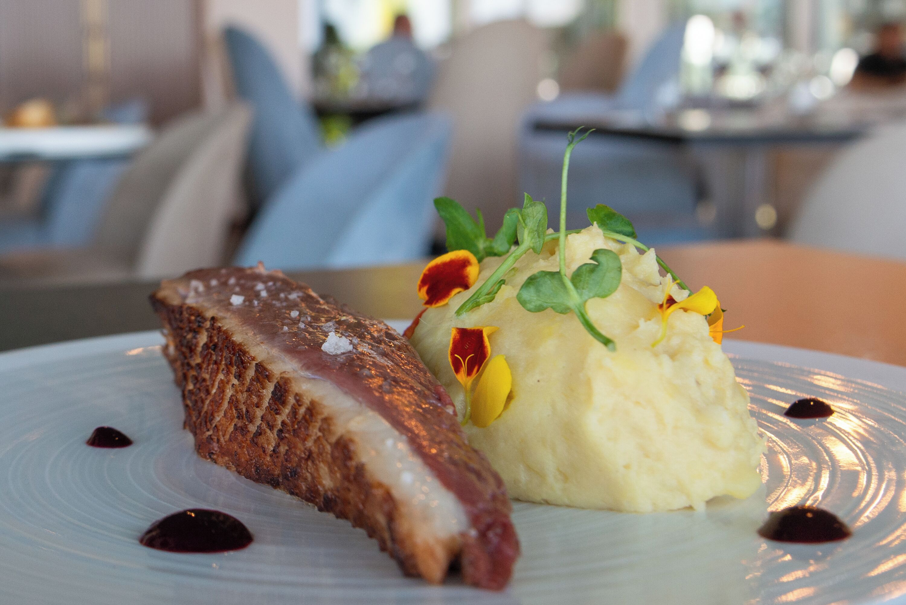 A plate with a piece of meat and mashed potatoes garnished with greens and flowers.