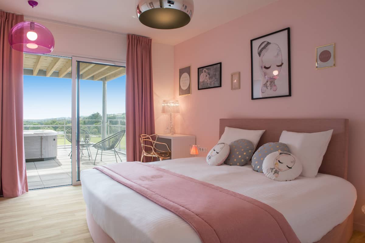 Een slaapkamer met een groot bed, een bureau en een stoel. Er is een balkon met een jacuzzi en uitzicht op het landschap.