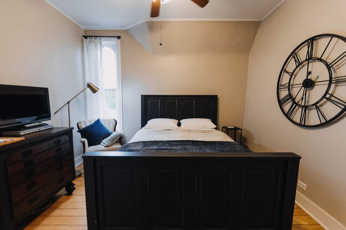 A bedroom with a bed, a dresser, a TV, a clock, and a window with curtains.