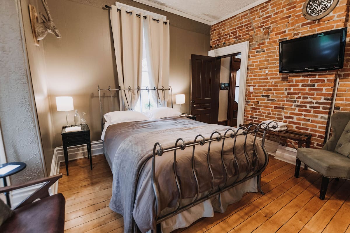 A hotel room with a bed, a chair, a TV, a lamp, and a brick wall.