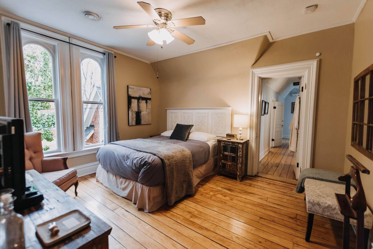 A bedroom with a large bed, wooden flooring, a ceiling fan, and a view of the outside through a window.