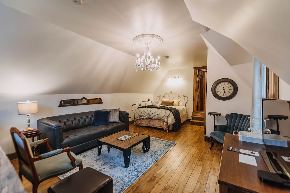 A spacious loft bedroom with a large bed, a sitting area with a sofa and armchairs, a wooden coffee table, and a chandelier.