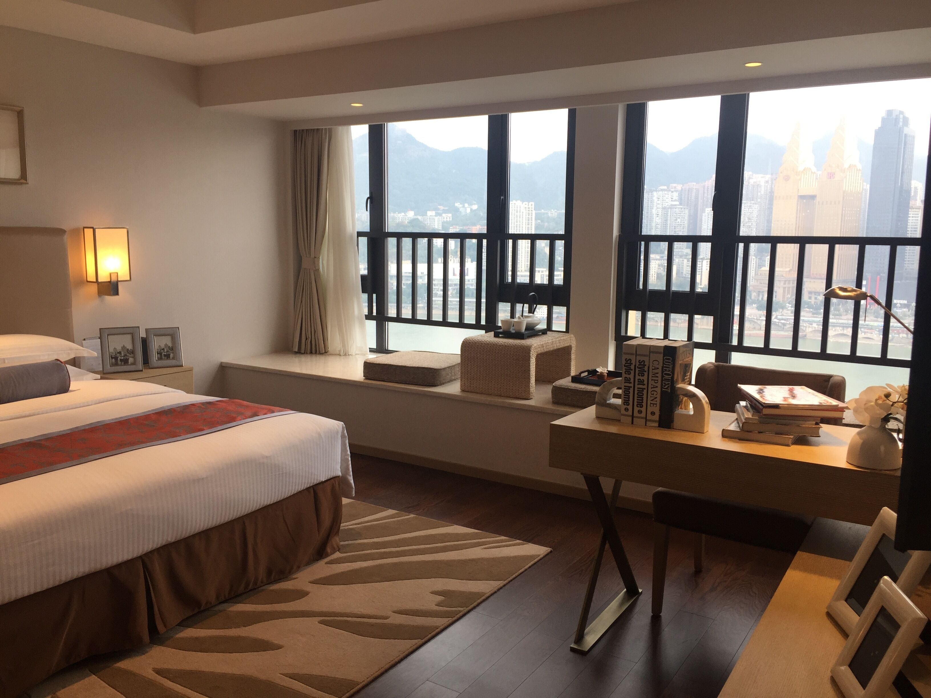 Habitación de hotel con una cama grande, un escritorio con libros, vistas a un paisaje urbano a través de amplios ventanales y un balcón con barandilla.