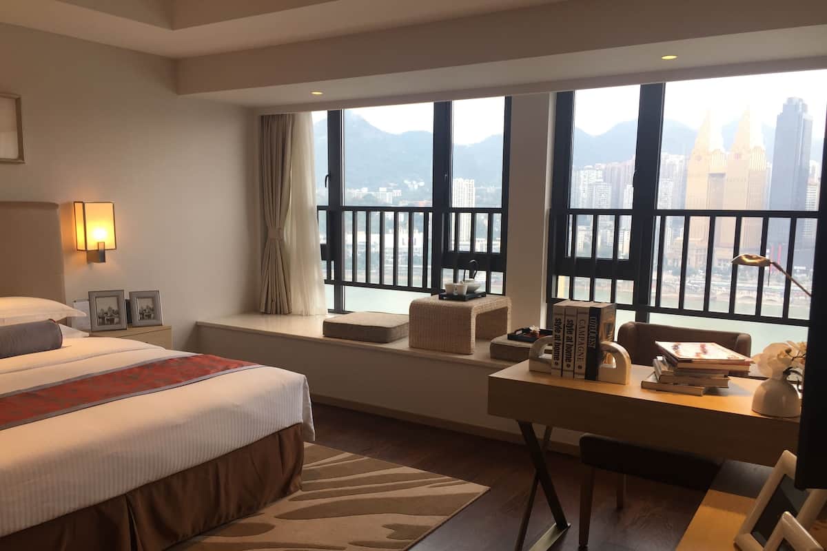 A hotel room with a large bed, a desk with books, a view of a cityscape through large windows, and a balcony with a railing.