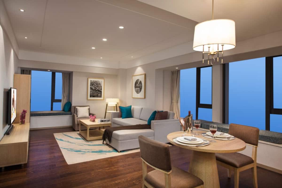 A modern hotel room with a sofa, dining table, and large windows offering a city view.