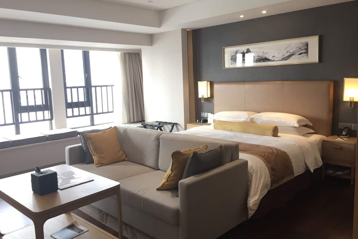 A hotel room with a bed, a sofa, a small table, a nightstand, and a window with a view of a balcony.