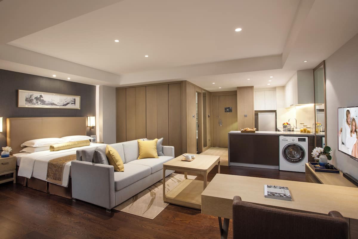 A modern hotel room with a sofa, a small table, a washing machine, and a kitchen area.