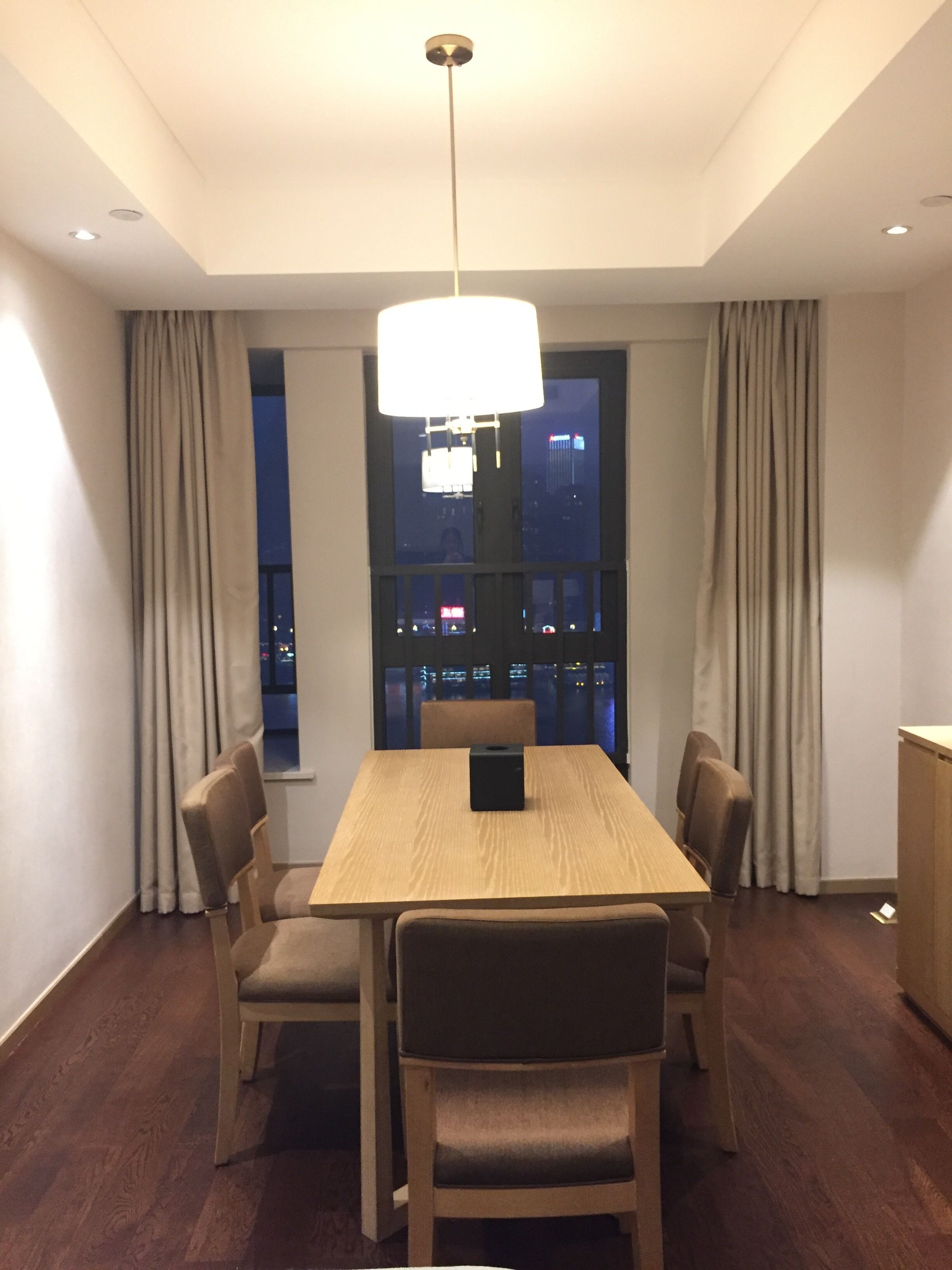 Un comedor moderno con una mesa de madera, cuatro sillas, una araña y vistas de la ciudad iluminada por la noche a través de amplios ventanales.