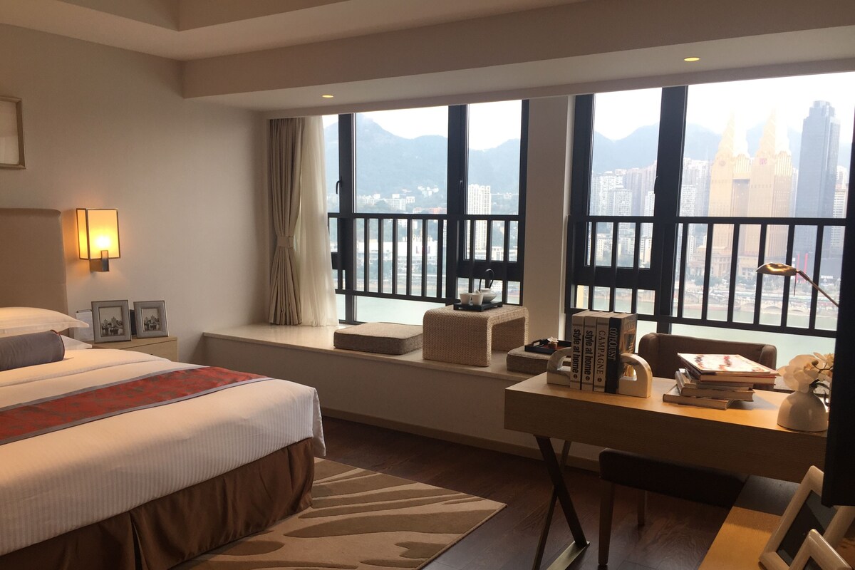 Habitación de hotel con una cama grande, un escritorio con libros, vistas a un paisaje urbano a través de amplios ventanales y un balcón con barandilla.