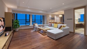 A spacious living room with a large sofa, a coffee table, and a television. There is a balcony visible through the large windows.