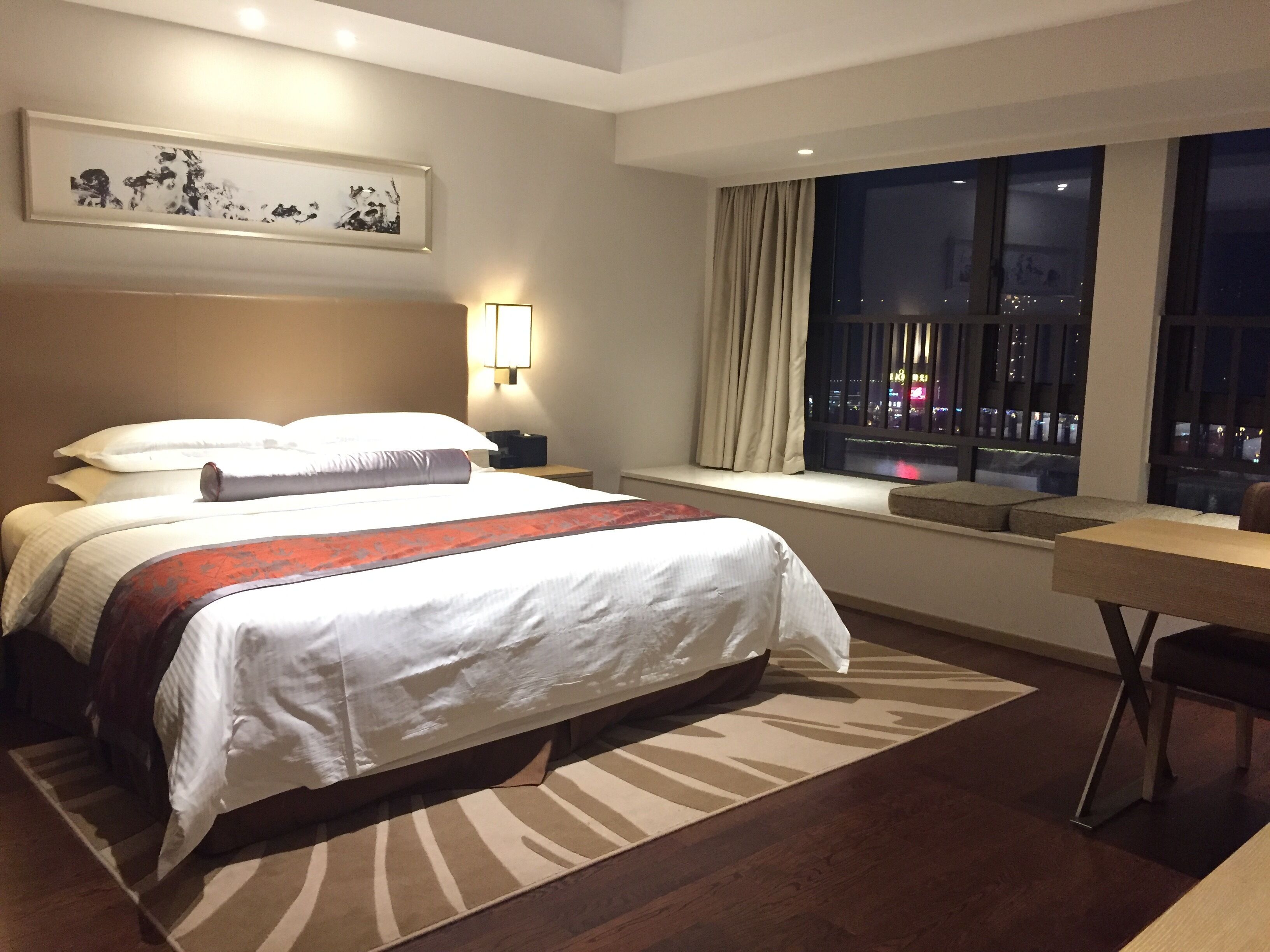 Une chambre d’hôtel avec un grand lit, un bureau, une fenêtre donnant sur la ville et une œuvre d’art encadrée au mur.