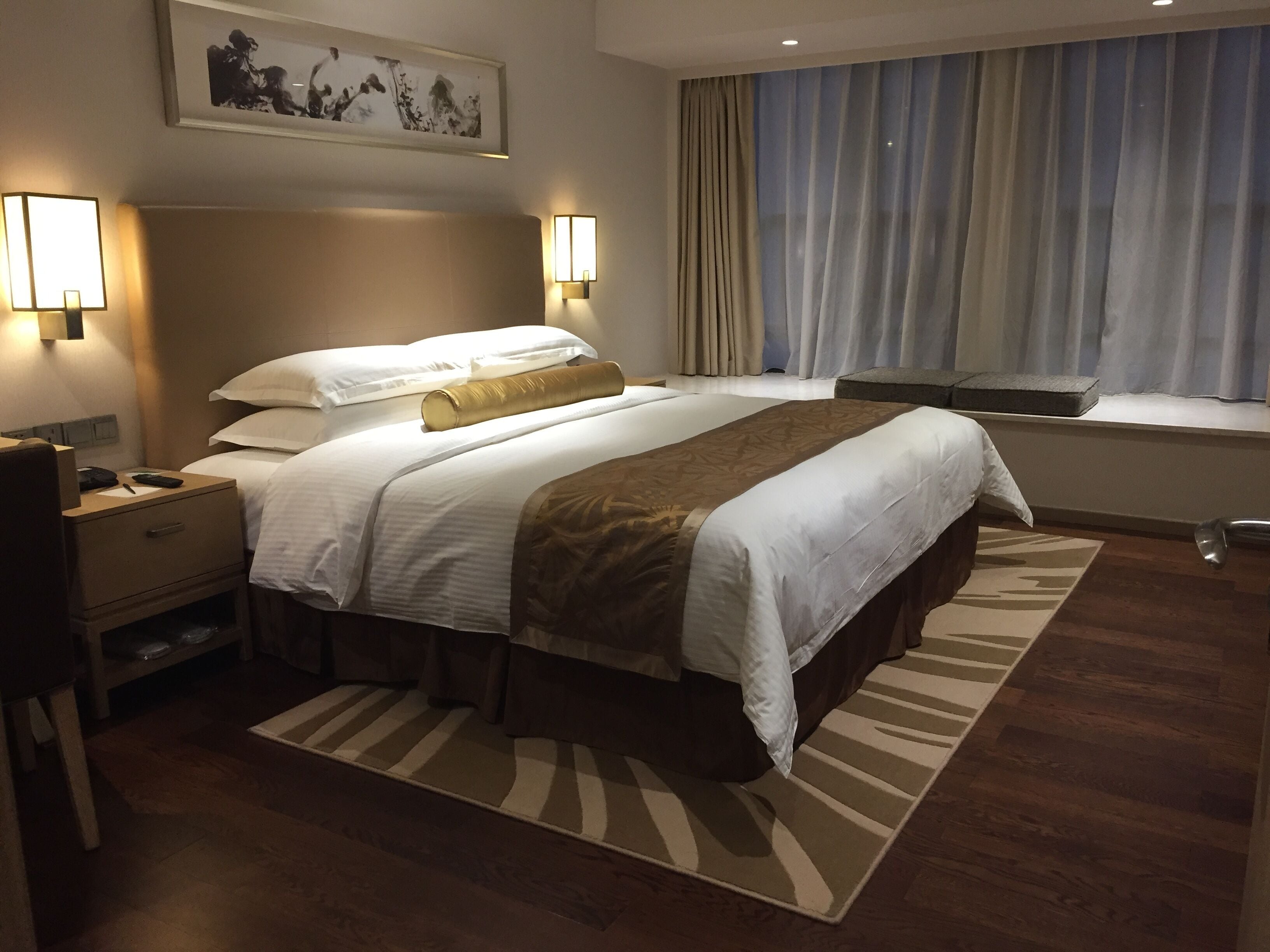 Une chambre d’hôtel avec un grand lit, des lampes de chevet, une table de chevet, une fenêtre avec des rideaux et un tableau encadré au mur.