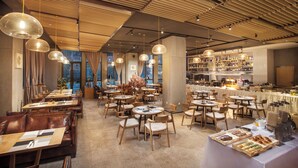 A spacious restaurant with multiple dining tables and chairs, a bar area with shelves of bottles, and a large window providing a view of the city.