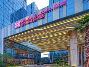 The exterior of the Hilton Garden Inn with a modern glass facade, a prominent entrance with a yellow-lit canopy, and a well-maintained garden area with trees and shrubs. - Hilton Garden Inn Ningbo (Ningbo)