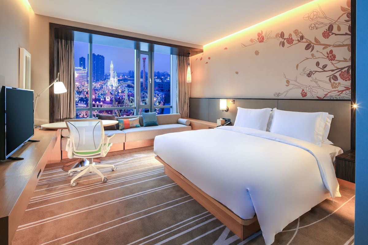 A hotel room with a large bed, a desk with a computer, a chair, and a view of a cityscape at night.