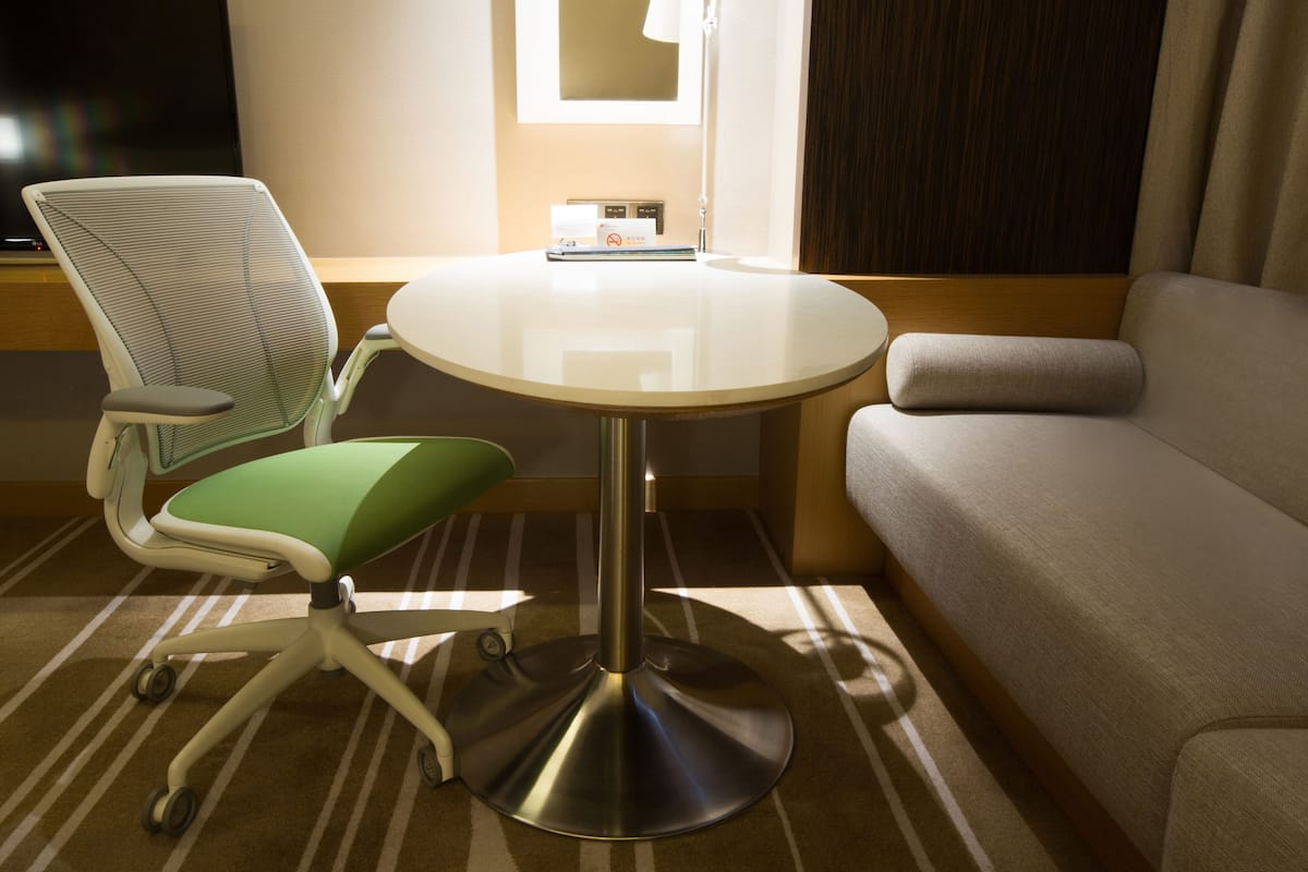 A hotel room with a round table, a green cushioned chair, a sofa, and a flat-screen TV.
