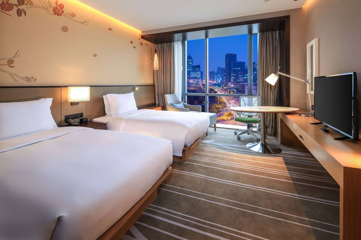 A hotel room with two beds, a desk with a TV, a chair, and a view of the city at night.