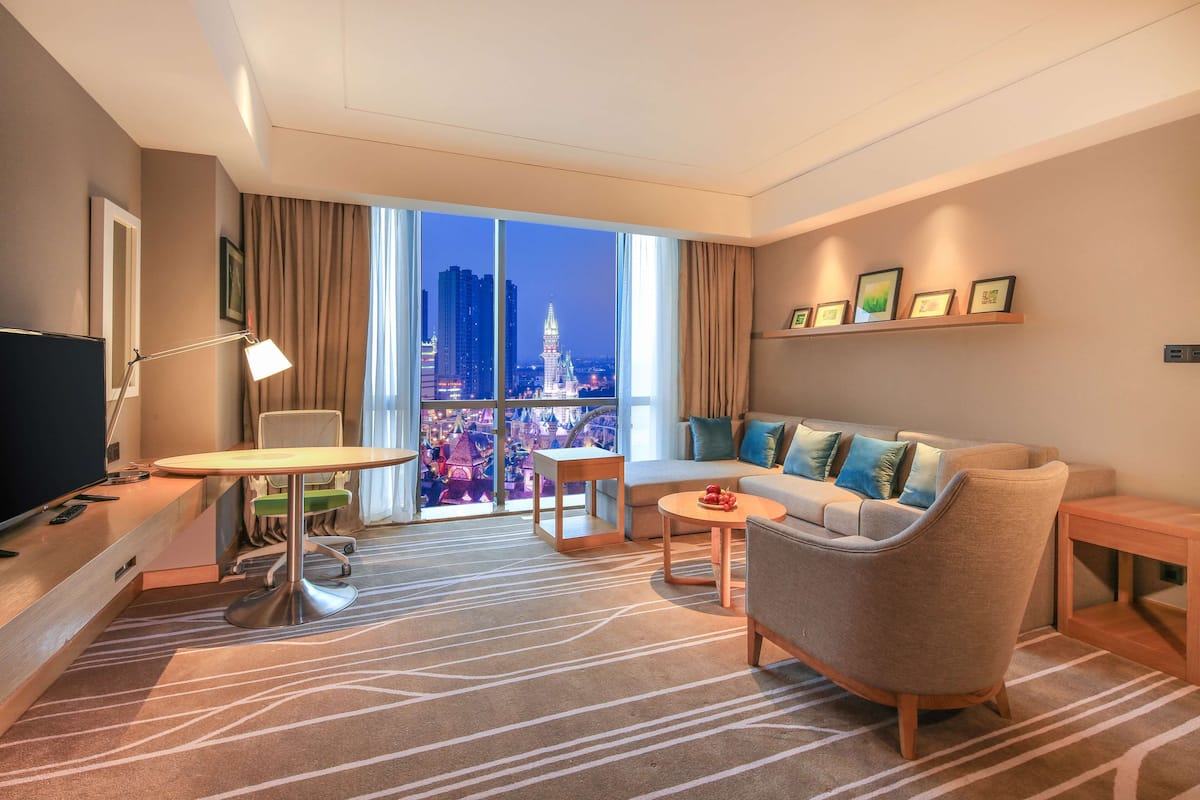 A modern hotel room with a large window offering a city view, a sofa, a small table, and a TV.