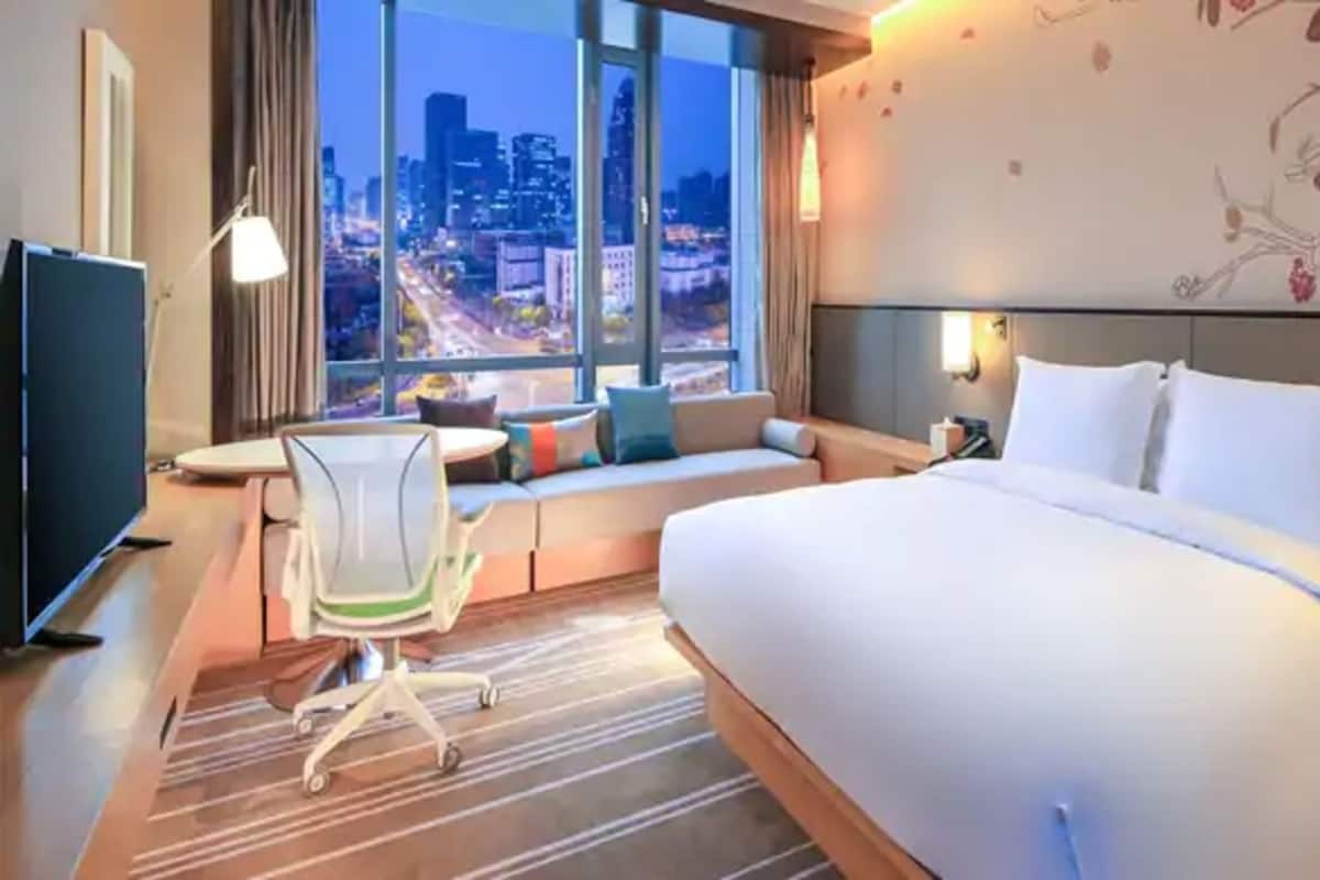 A hotel room with a large bed, a desk with a chair, a TV, and a view of the cityscape.