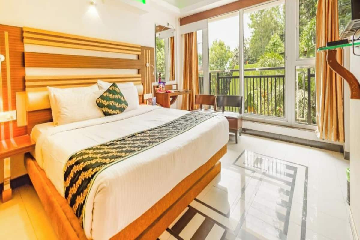 A hotel room with a large bed, a desk, a chair, and a balcony with a view of greenery.