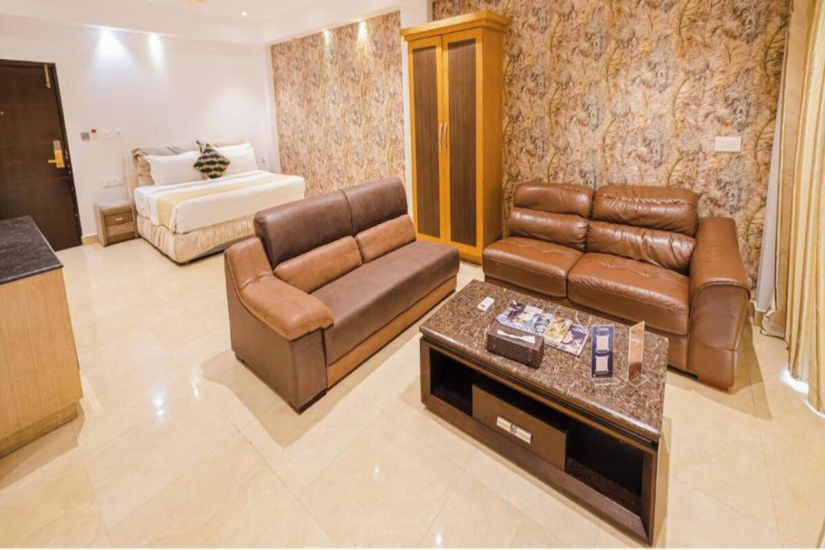 A spacious living room with a brown leather sofa, a matching armchair, and a coffee table with magazines. A wooden wardrobe and a bed with white bedding are also visible.