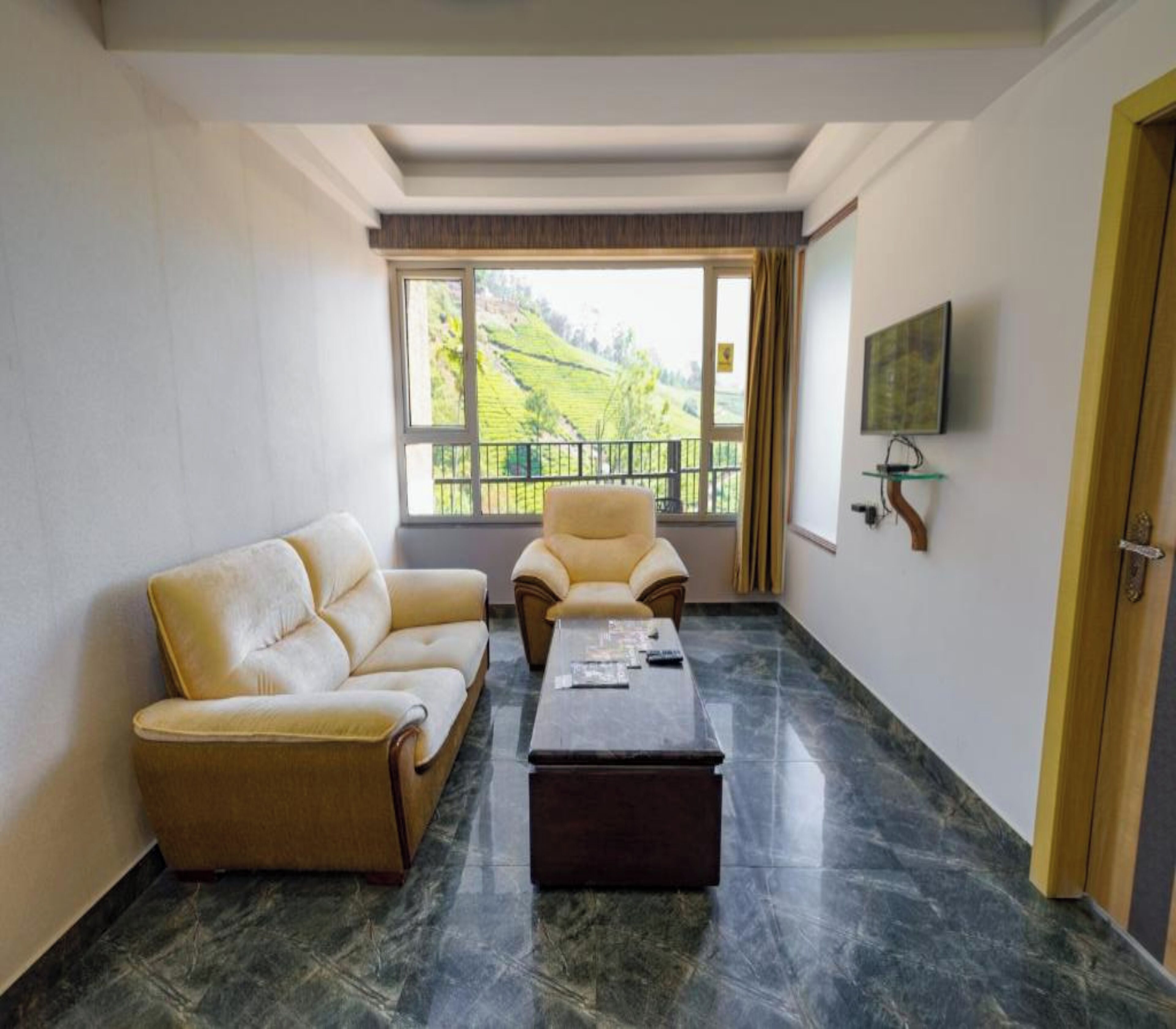 A living room with a sofa, armchair, and coffee table, a TV mounted on the wall, and a large window with a view of a green landscape.