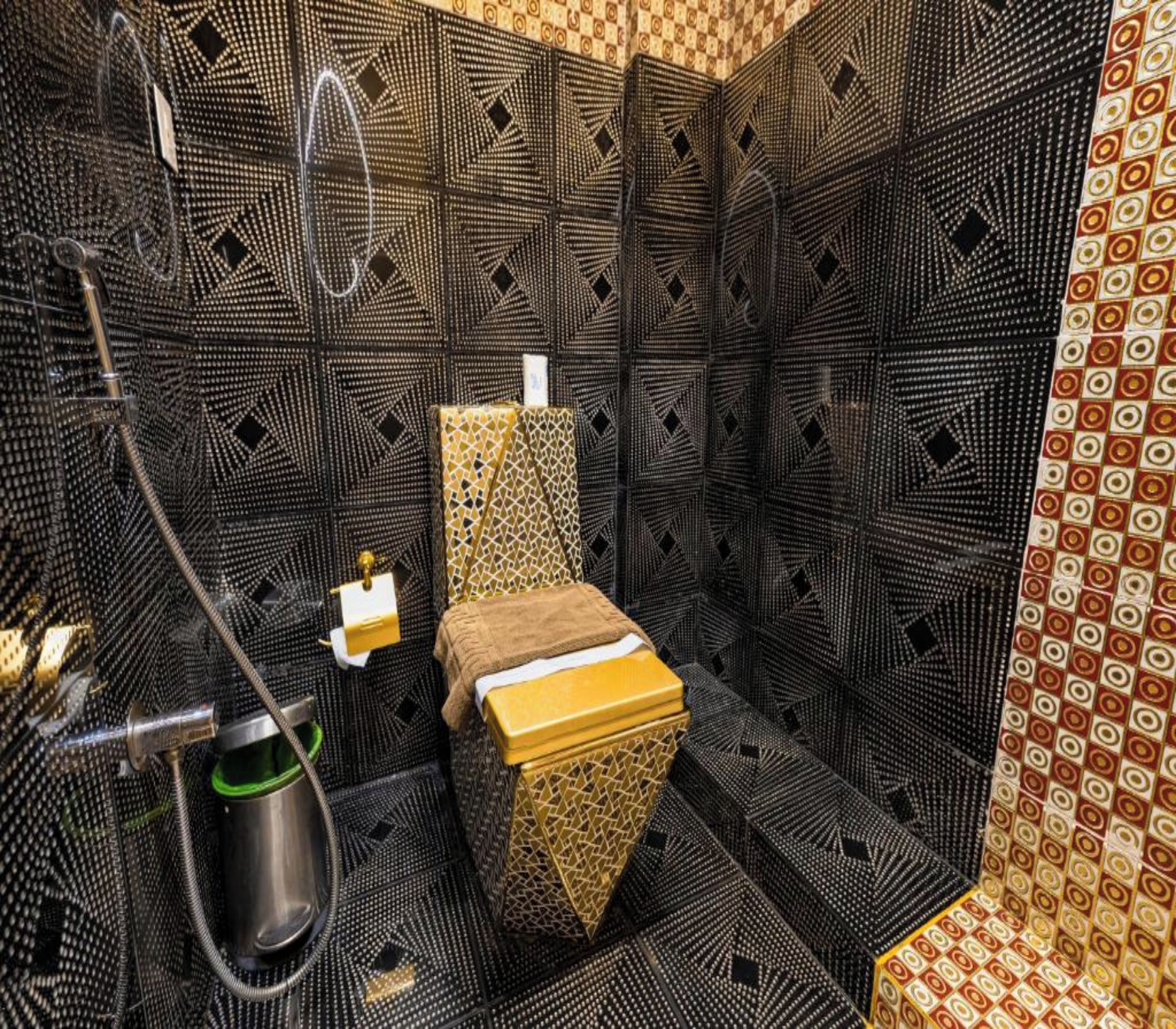 A small, enclosed bathroom with a patterned floor and walls, a toilet, a sink, and a handheld showerhead.