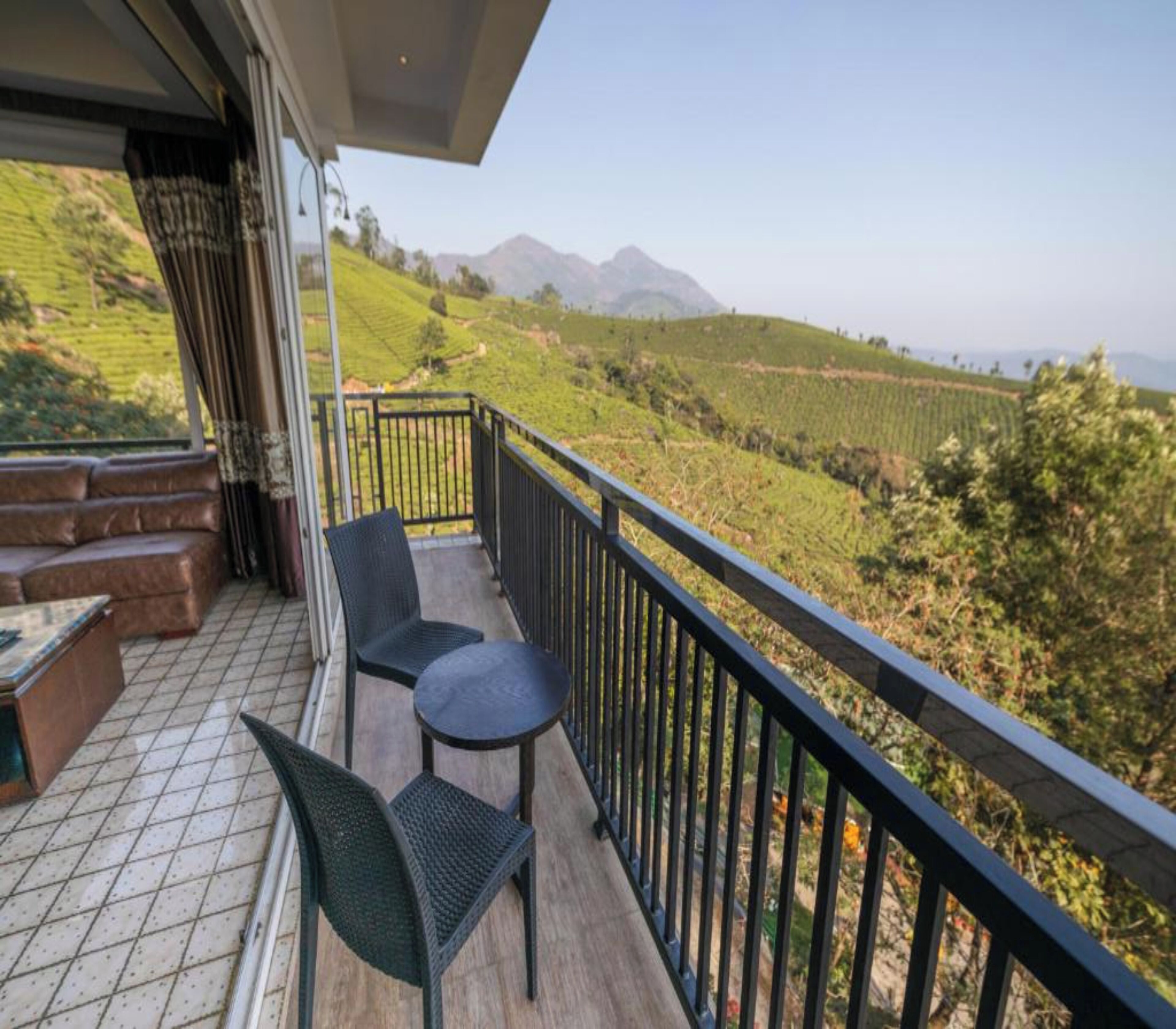 A balcony with two chairs and a small table, overlooking a hilly landscape with tea plantations.