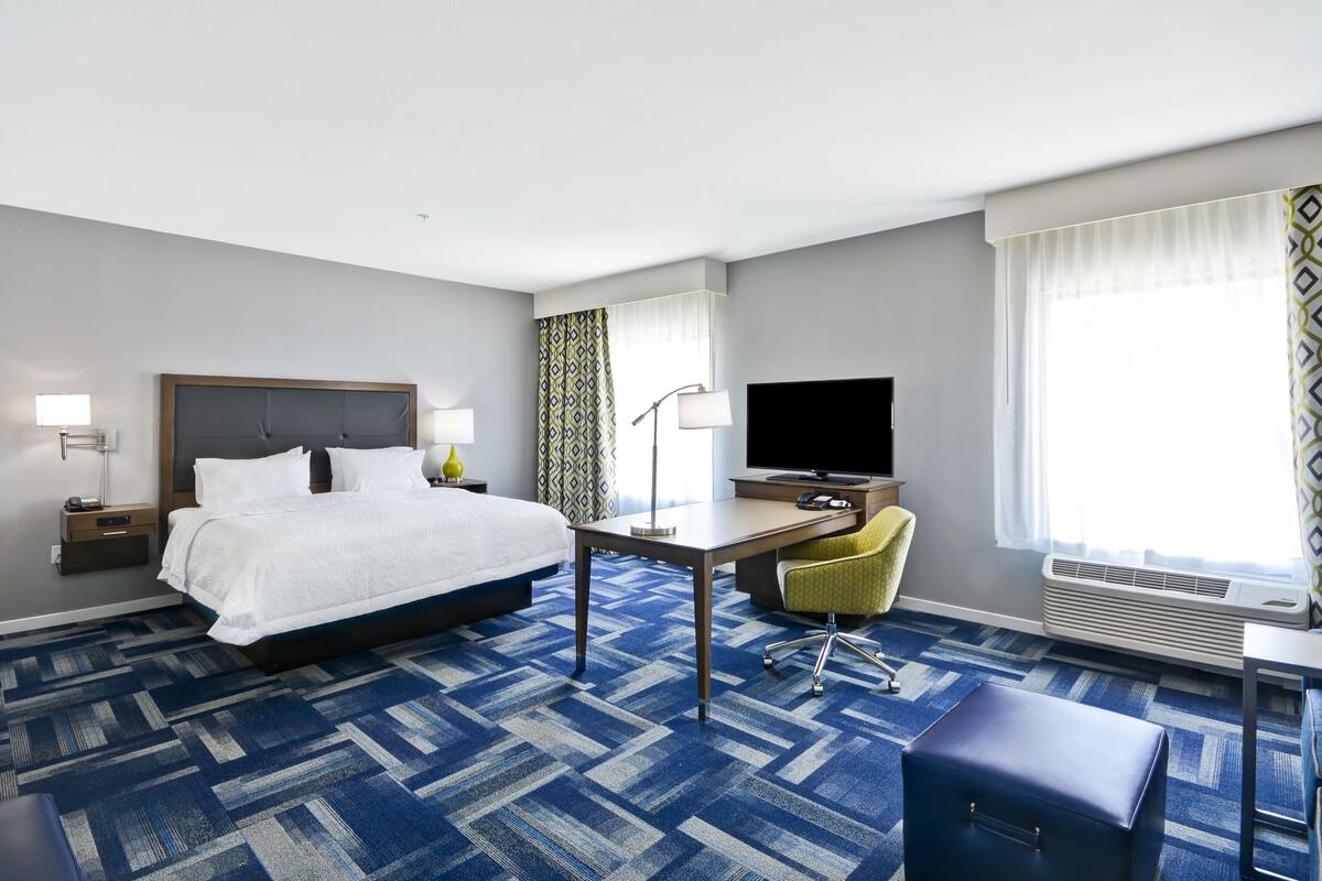 A hotel room with a large bed, a desk with a chair, a television, and an air conditioning unit.