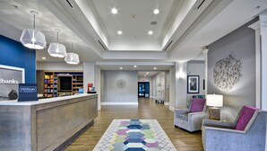 A hotel lobby with a reception desk, a seating area with two armchairs, a patterned rug, and a geometric wall art piece. - Hampton Inn Kyle (Kyle)