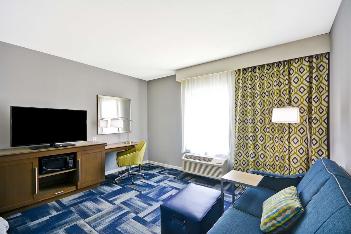 A hotel room with a flat-screen TV, a desk with a chair, a sofa with a pillow, and patterned curtains.