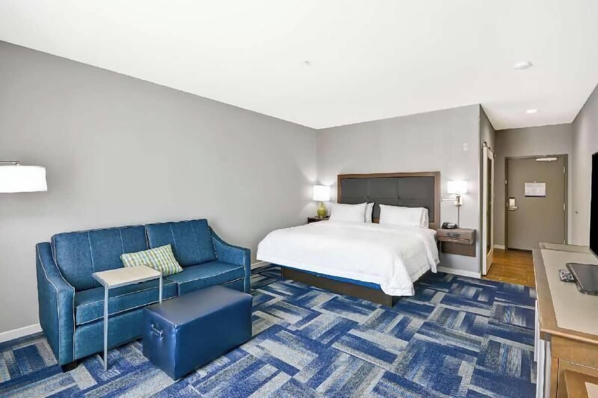 A hotel room with a bed, a blue sofa, a small table, a blue ottoman, and a desk with a chair.