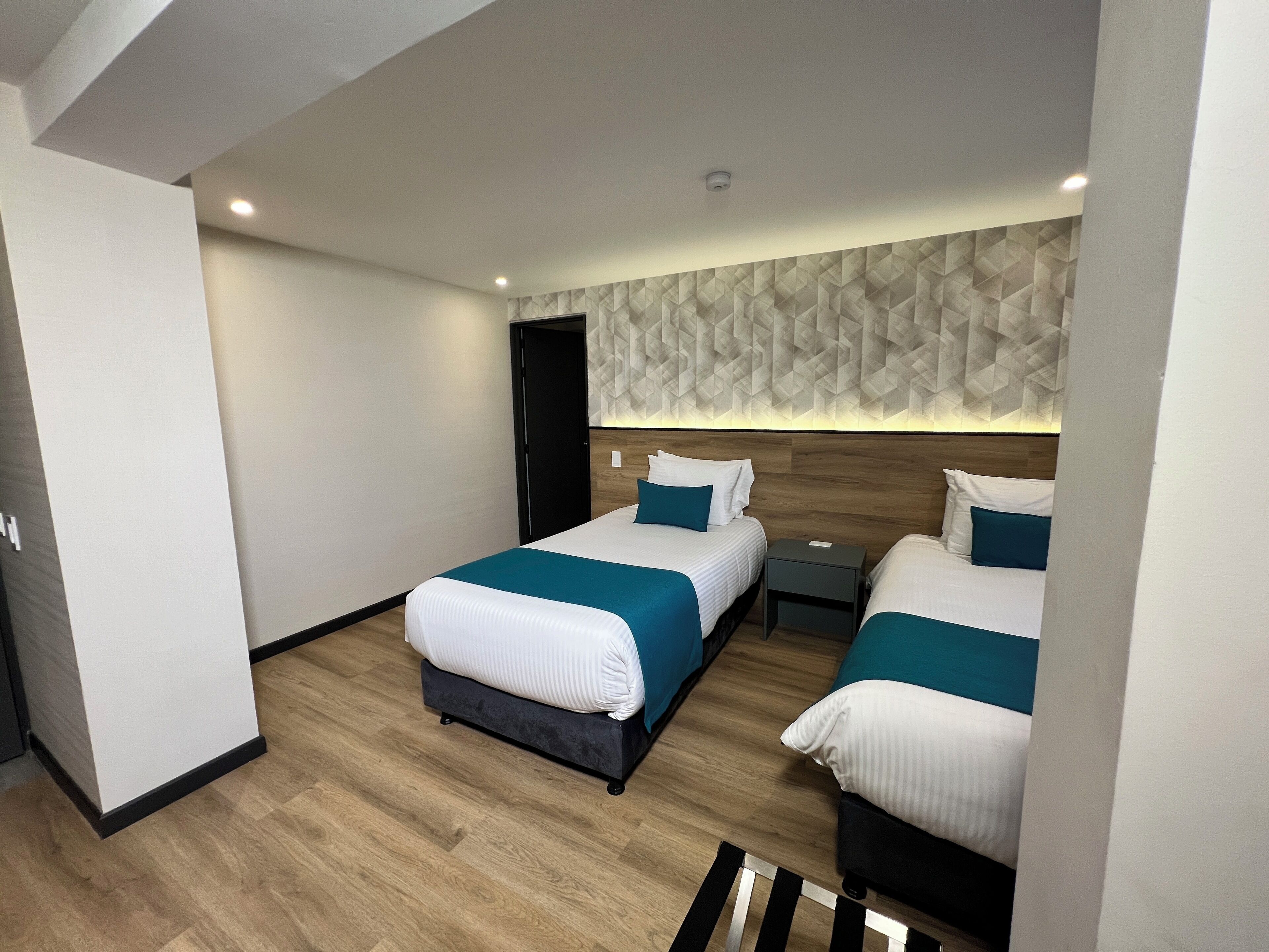 Una habitación de hotel moderna con dos camas, piso de madera, pared con estampado geométrico y una mesita de noche pequeña.