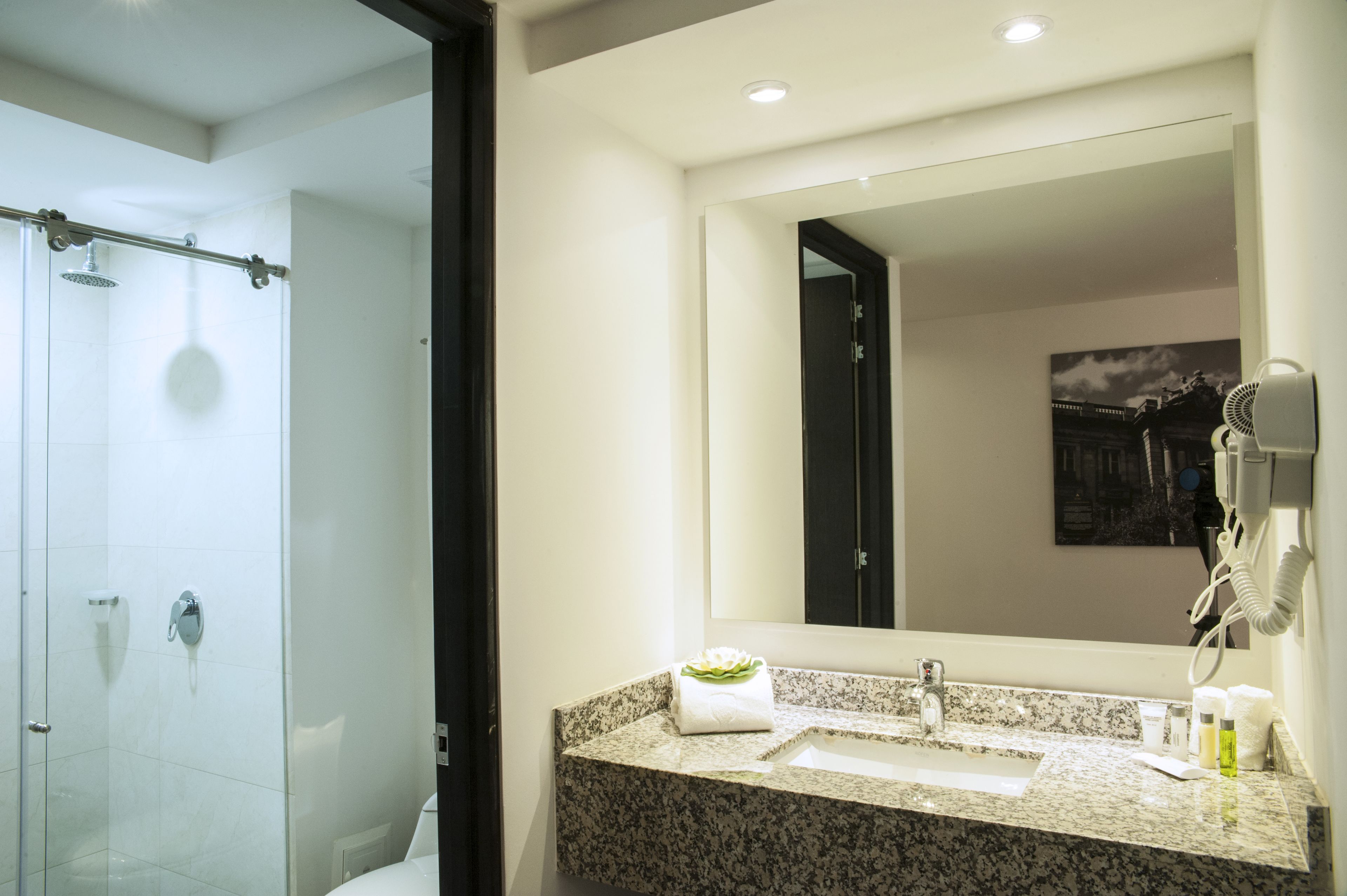 Un baño moderno con ducha de vidrio, encimera de granito, un espejo grande y un secador de pelo montado en la pared.