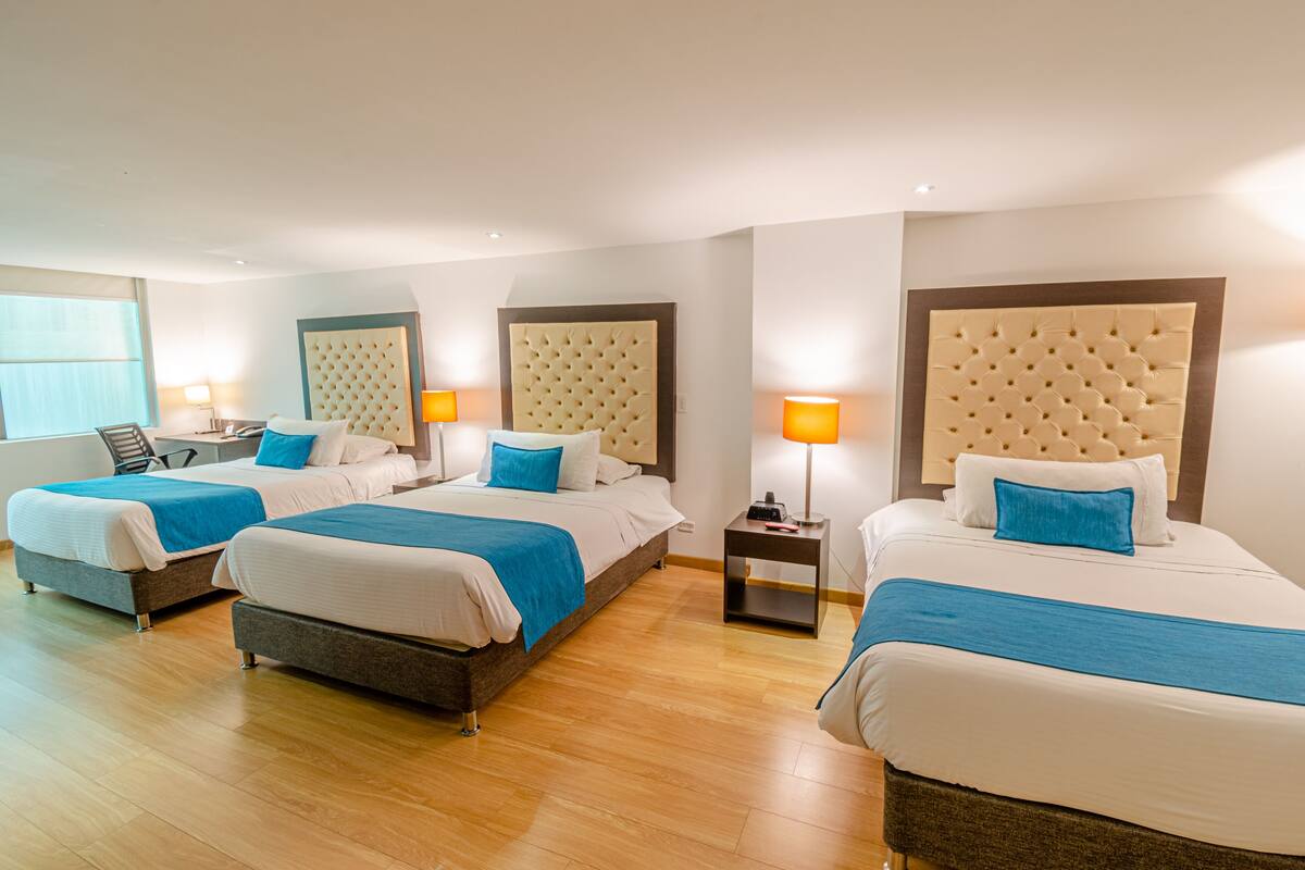 A hotel room with three beds, each with a tufted headboard, a bedside table with a lamp, and a window with blinds.