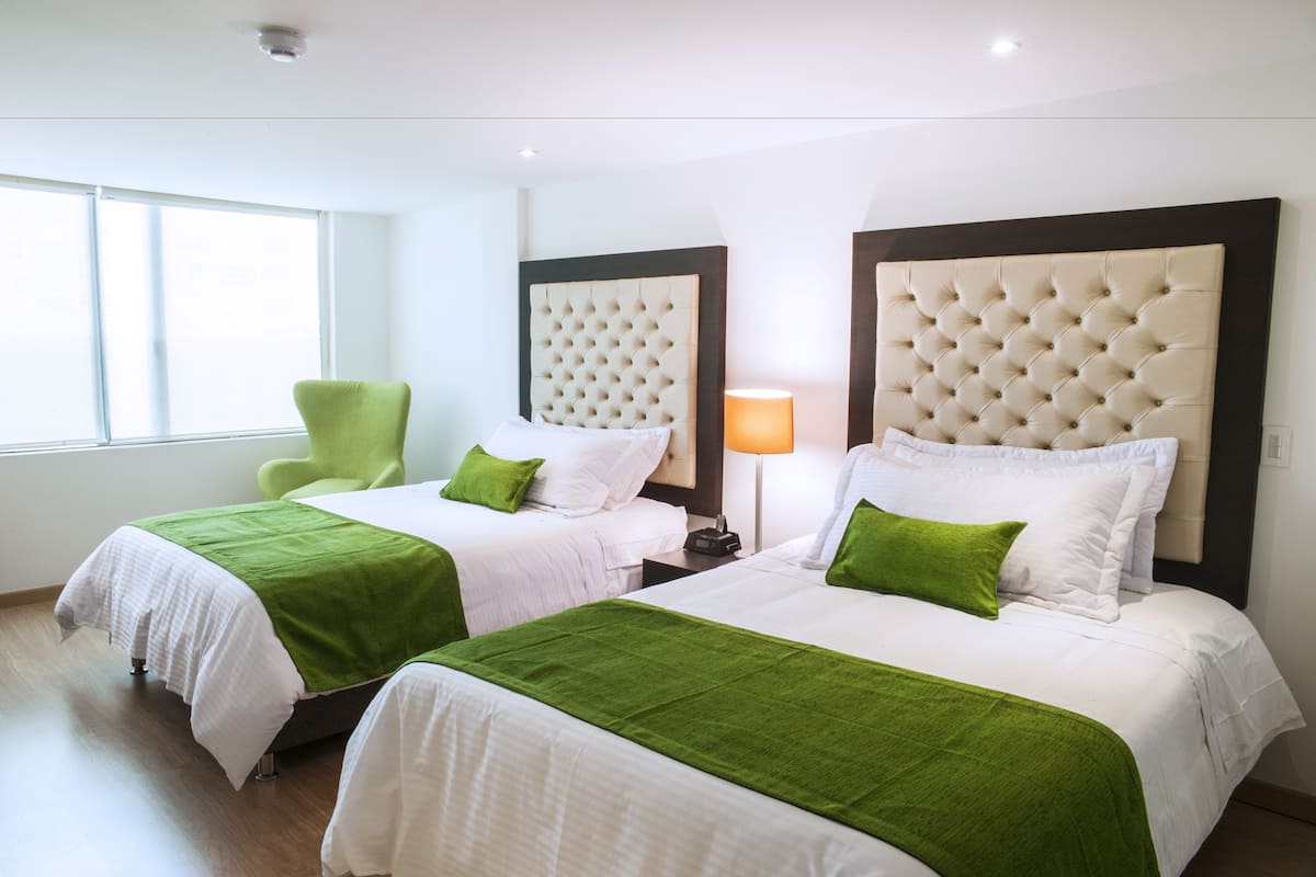 A hotel room with two beds, green and white bedding, a green chair, and a nightstand with a lamp.