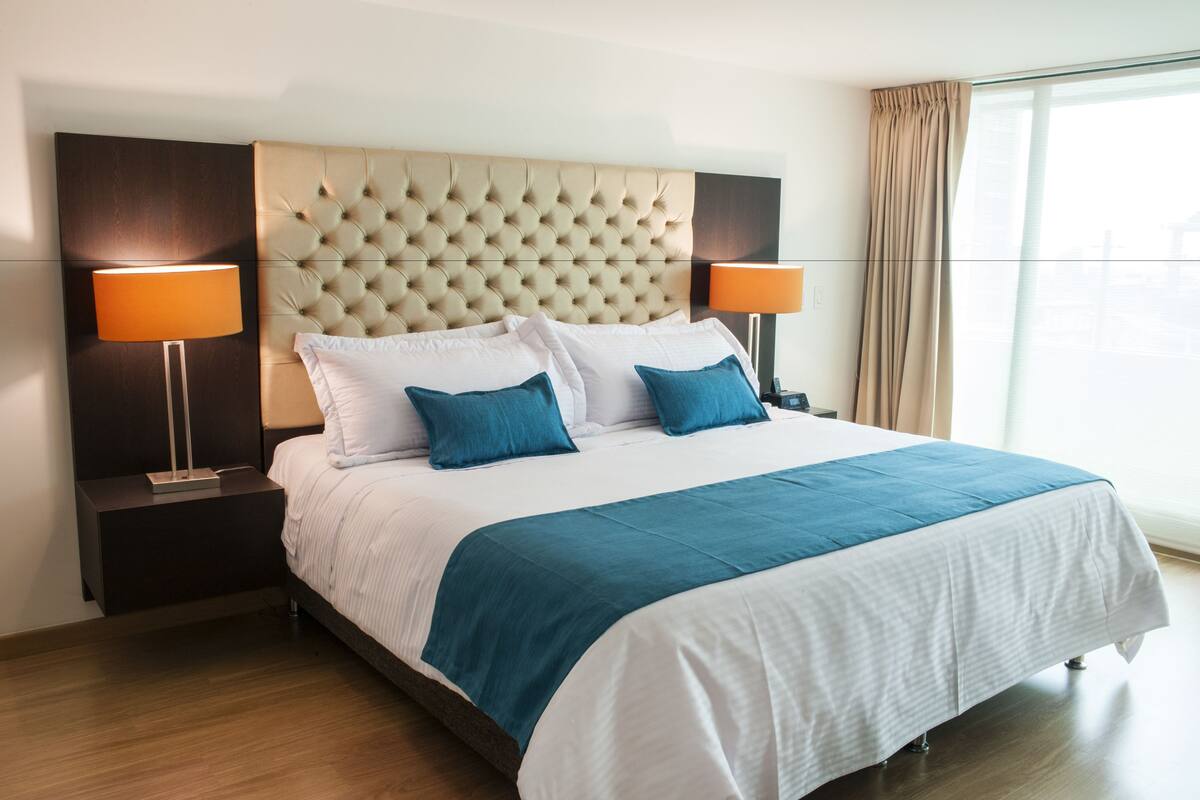 A hotel room with a large bed, two bedside lamps, a tufted headboard, and a large window with curtains.