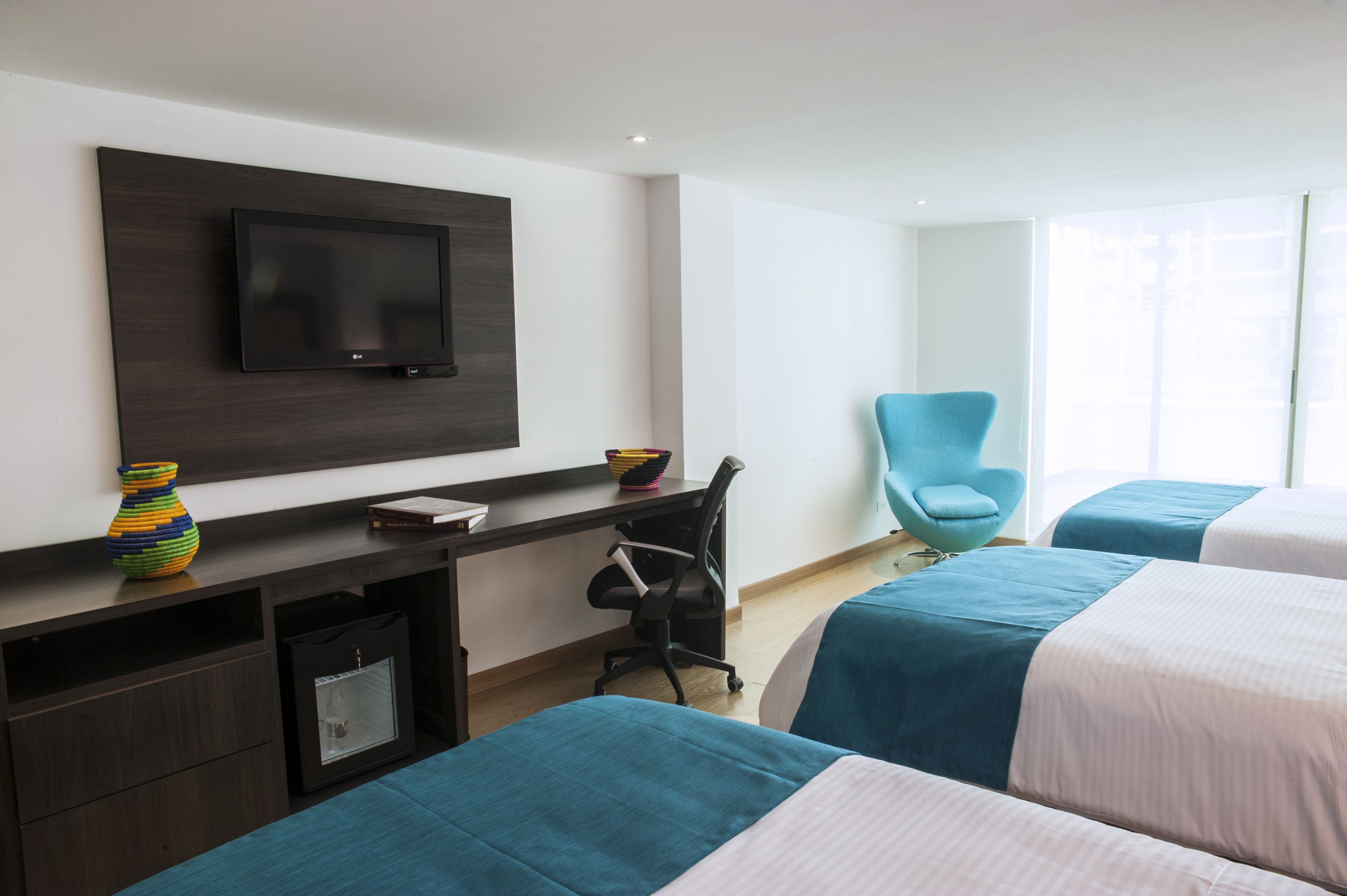 A hotel room with a flat-screen TV mounted on the wall, a desk with a chair, and two beds with teal bedding.