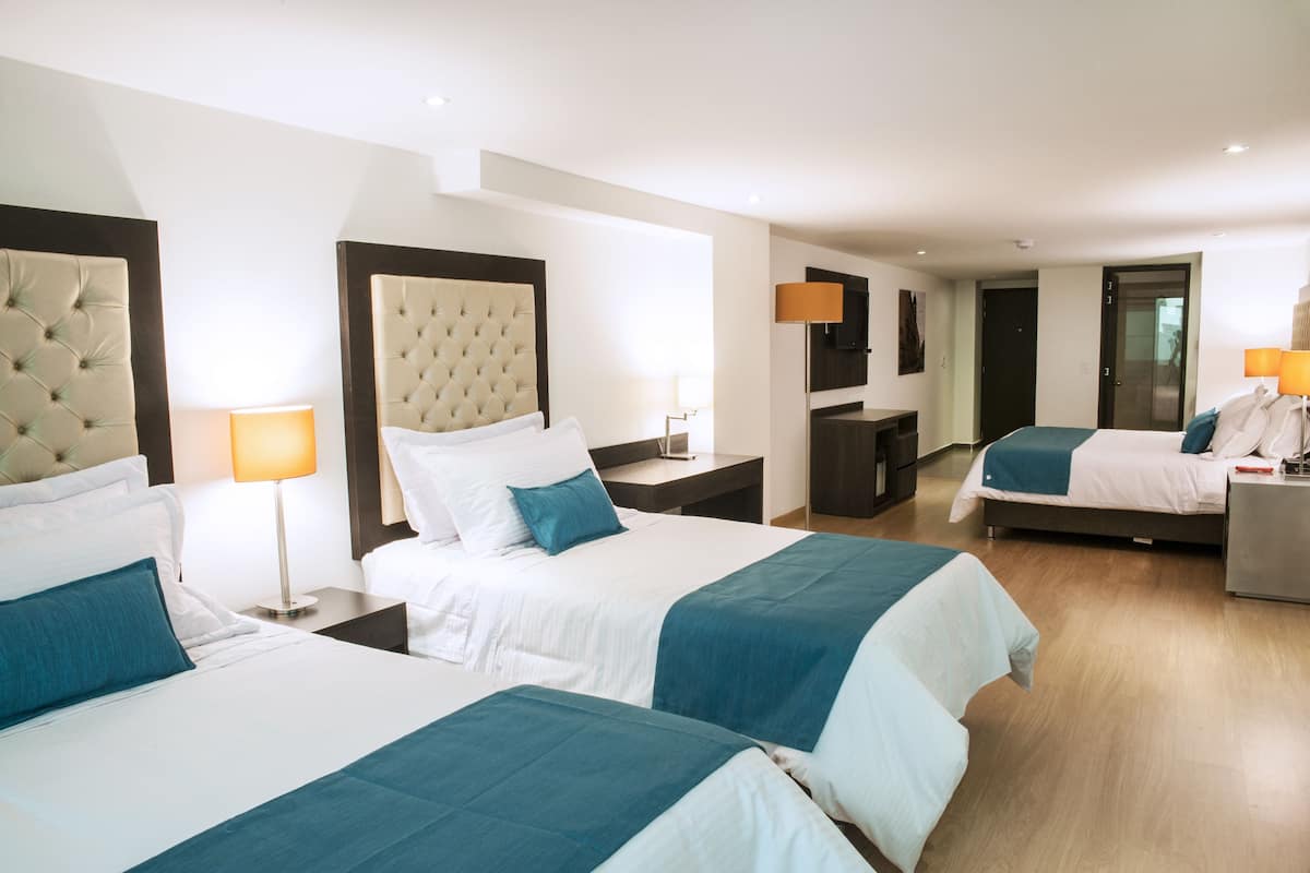 A hotel room with two beds, a large tufted headboard, a nightstand with a lamp, a desk with a chair, and a wardrobe.