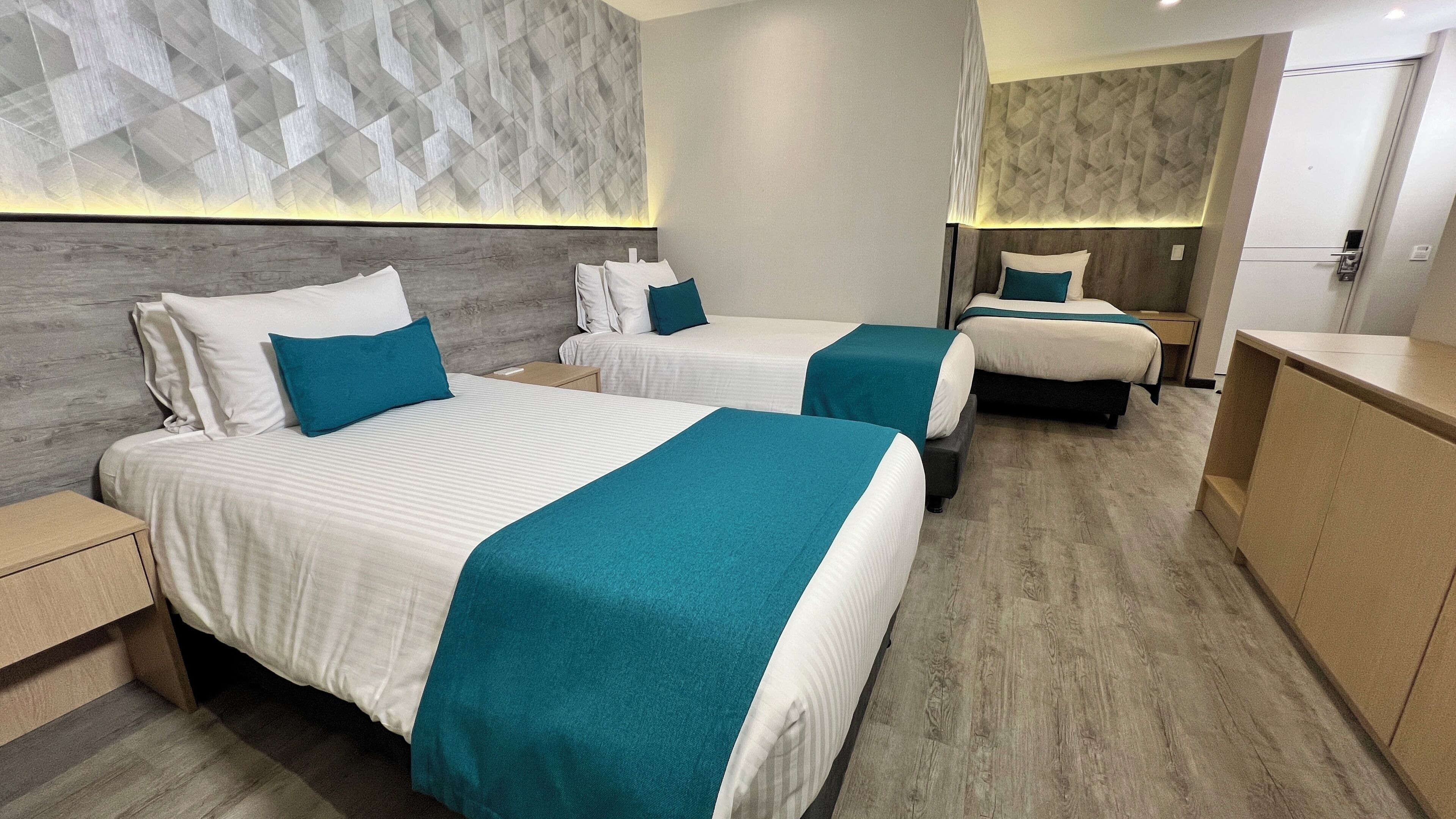 Habitación de hotel con dos camas, cada una con ropa de cama blanca y detalles en color verde azulado, cabeceros de madera y un diseño de pared moderno y geométrico.