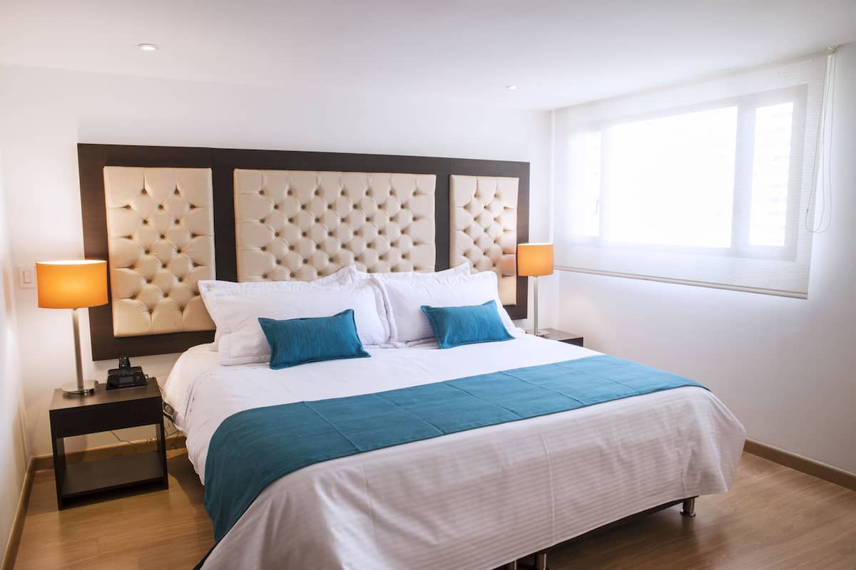 A neatly made bed with a tufted headboard, two bedside lamps, and a window with blinds.